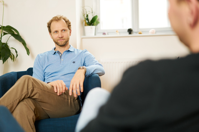 Blick über die Schulter einer Person im Gespräch mit Psychotherapeut Richard Stark, der dem Klienten in Jeans und blauem Hemd gegenüber sitzt.